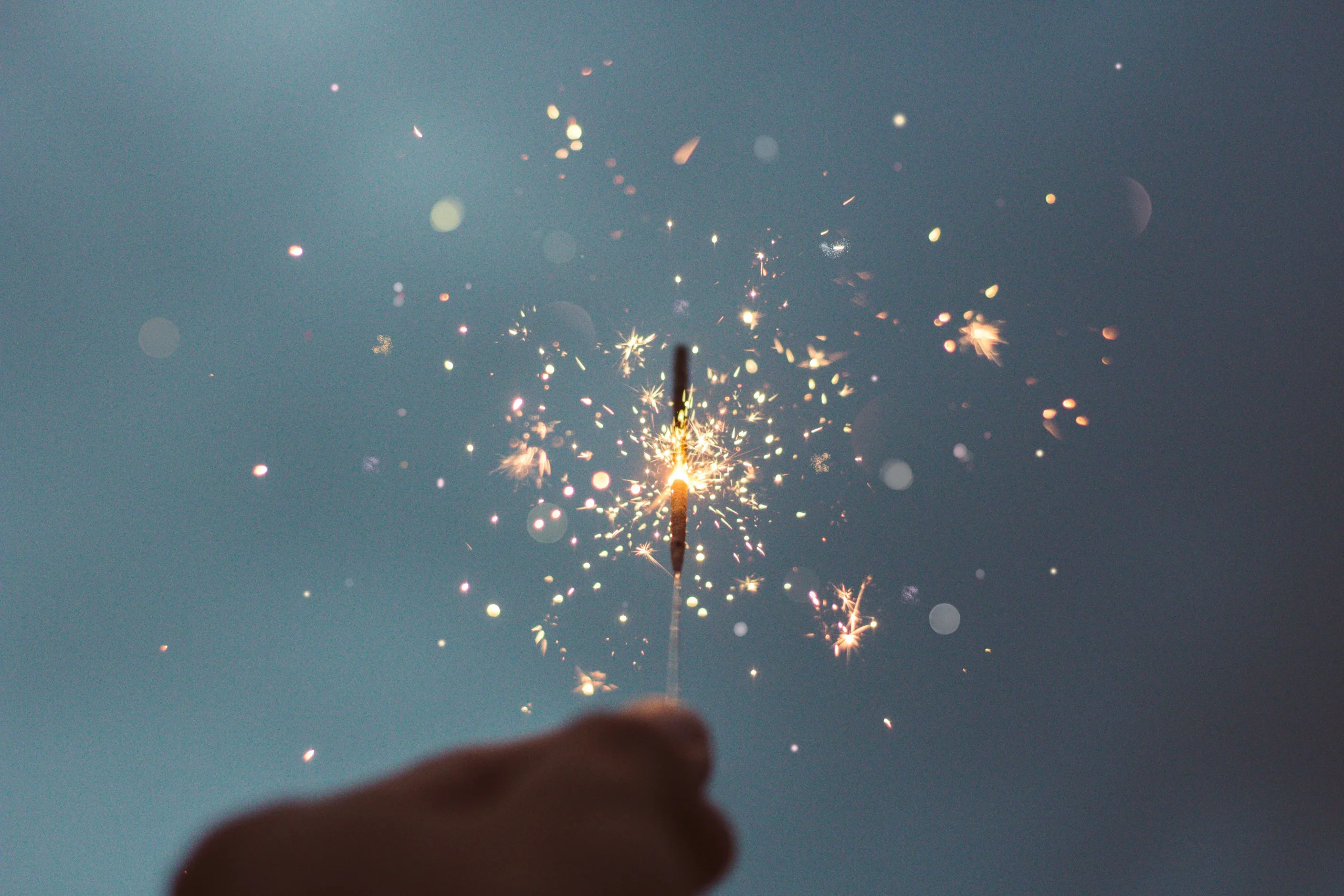 A hand holding a lit sparkler against a dark night sky, with bright, colorful sparks illuminating the scene