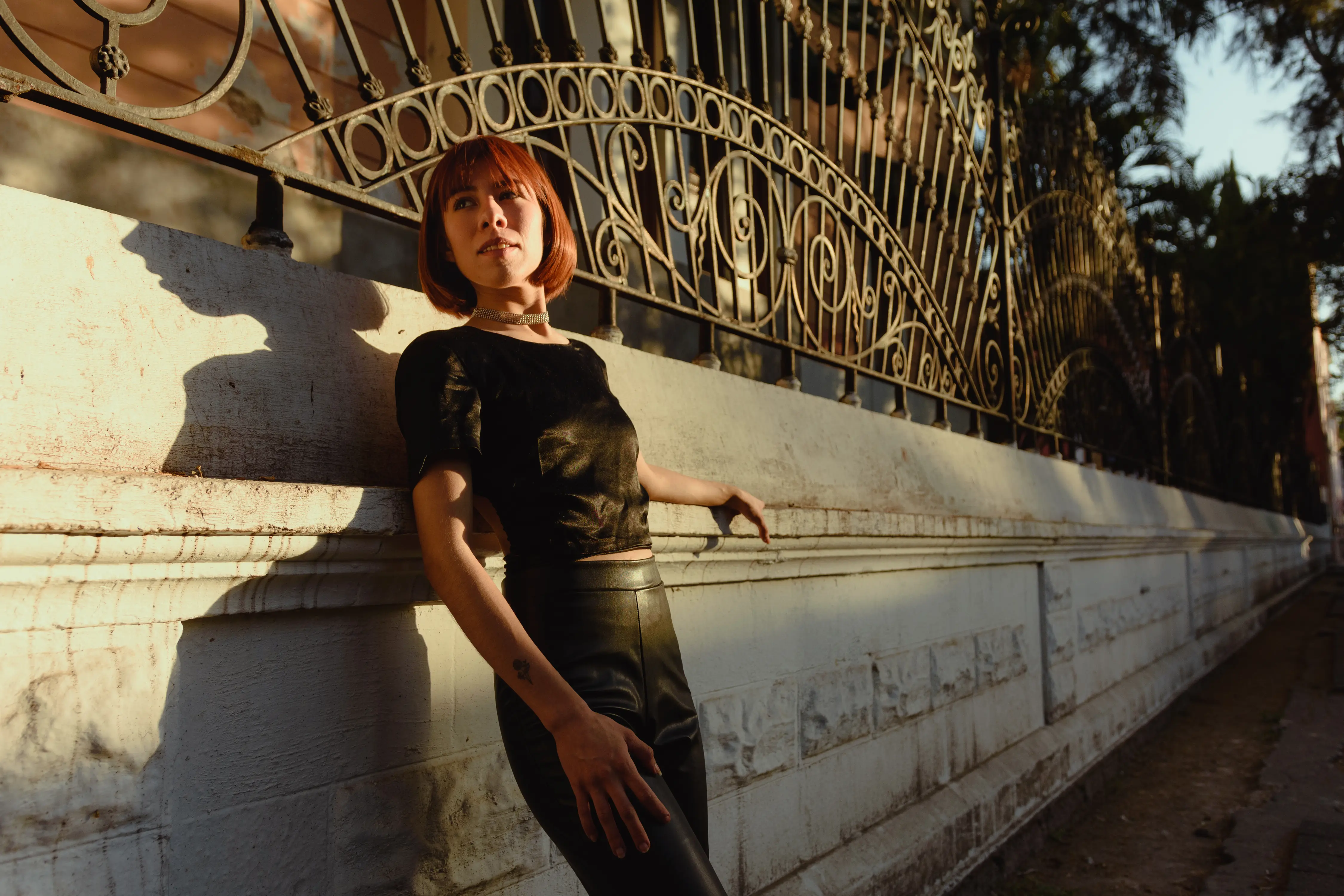 Full-body portrait of a woman with short auburn hair, leaning against an ornate iron fence at sunset, wearing a black outfit and a sparkling choker necklace.